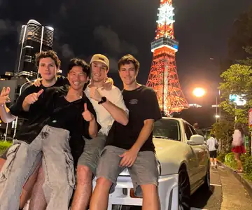 Un grupo sentado sobre un coche deportivo blanco, con la Torre de Tokio iluminada de noche