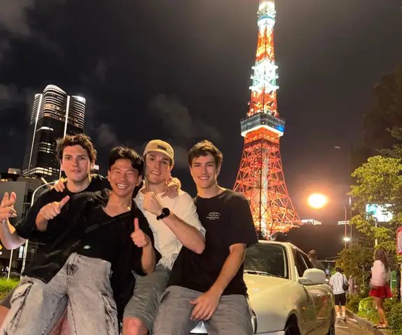Un grupo sentado sobre un coche deportivo blanco, con la Torre de Tokio iluminada de noche