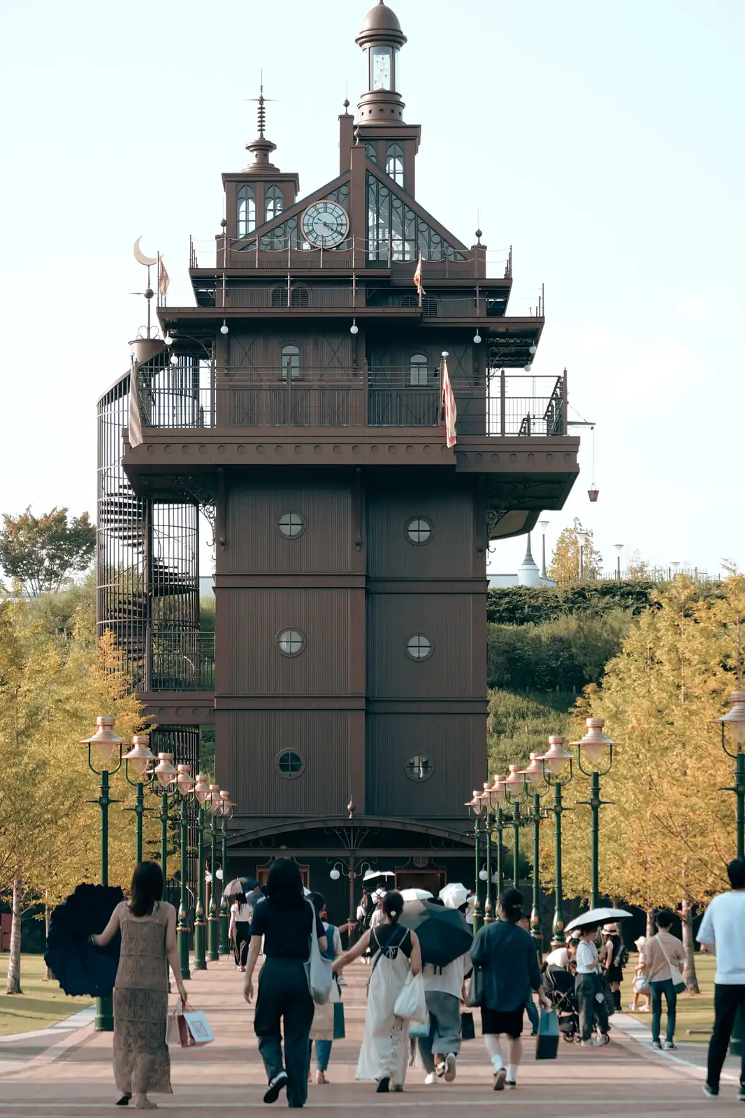 Torre de estilo antiguo con reloj al final de un paseo con farolas, en Ghibli Park